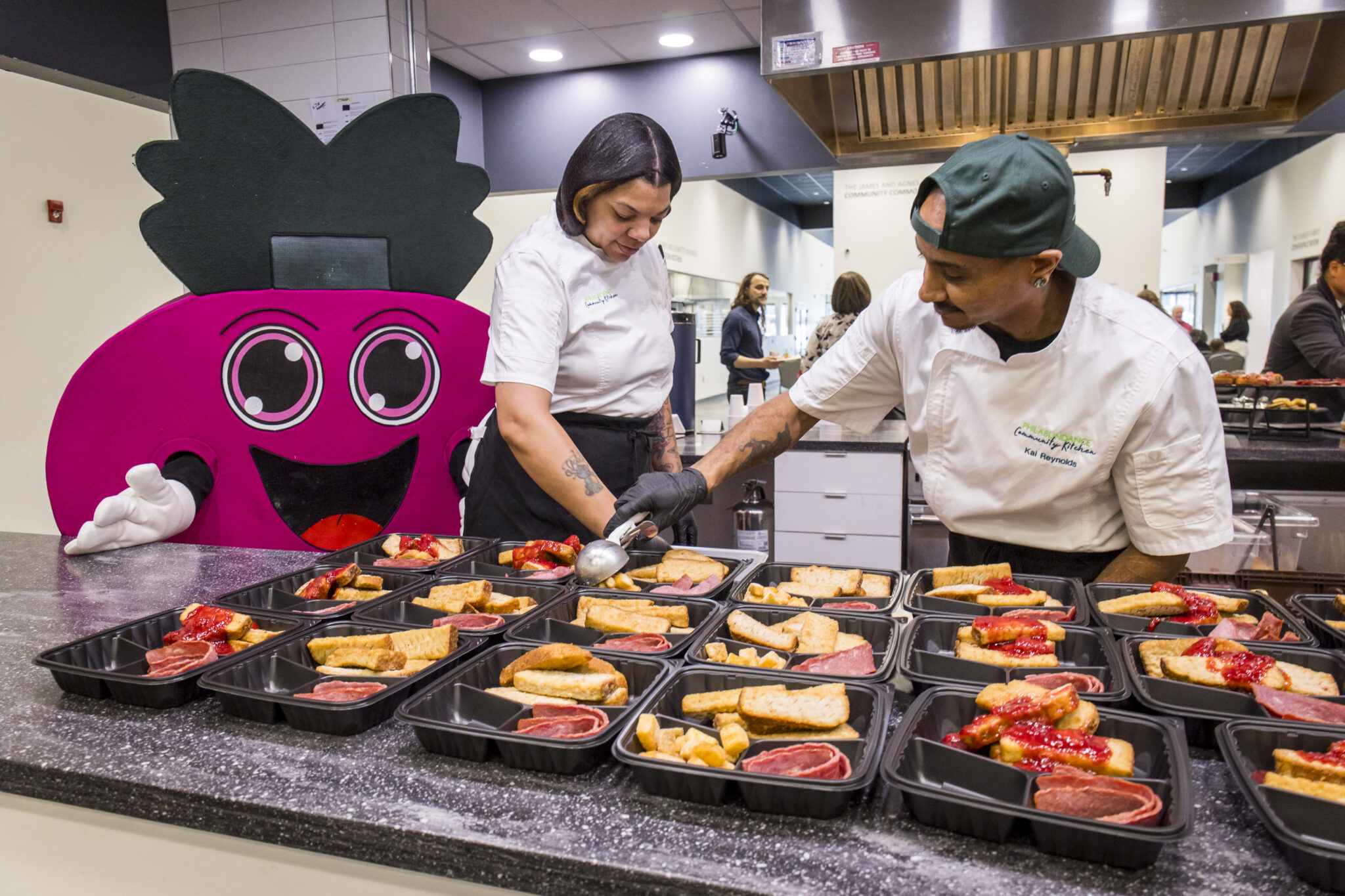 Two chefs in white coats prepare rows of black trays of meals while the Philabundance mascot which is a purple and black cartoonish beet looks on