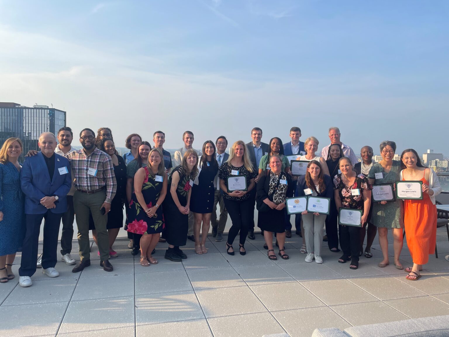 A large group of people stand on a roof deck holding their awards