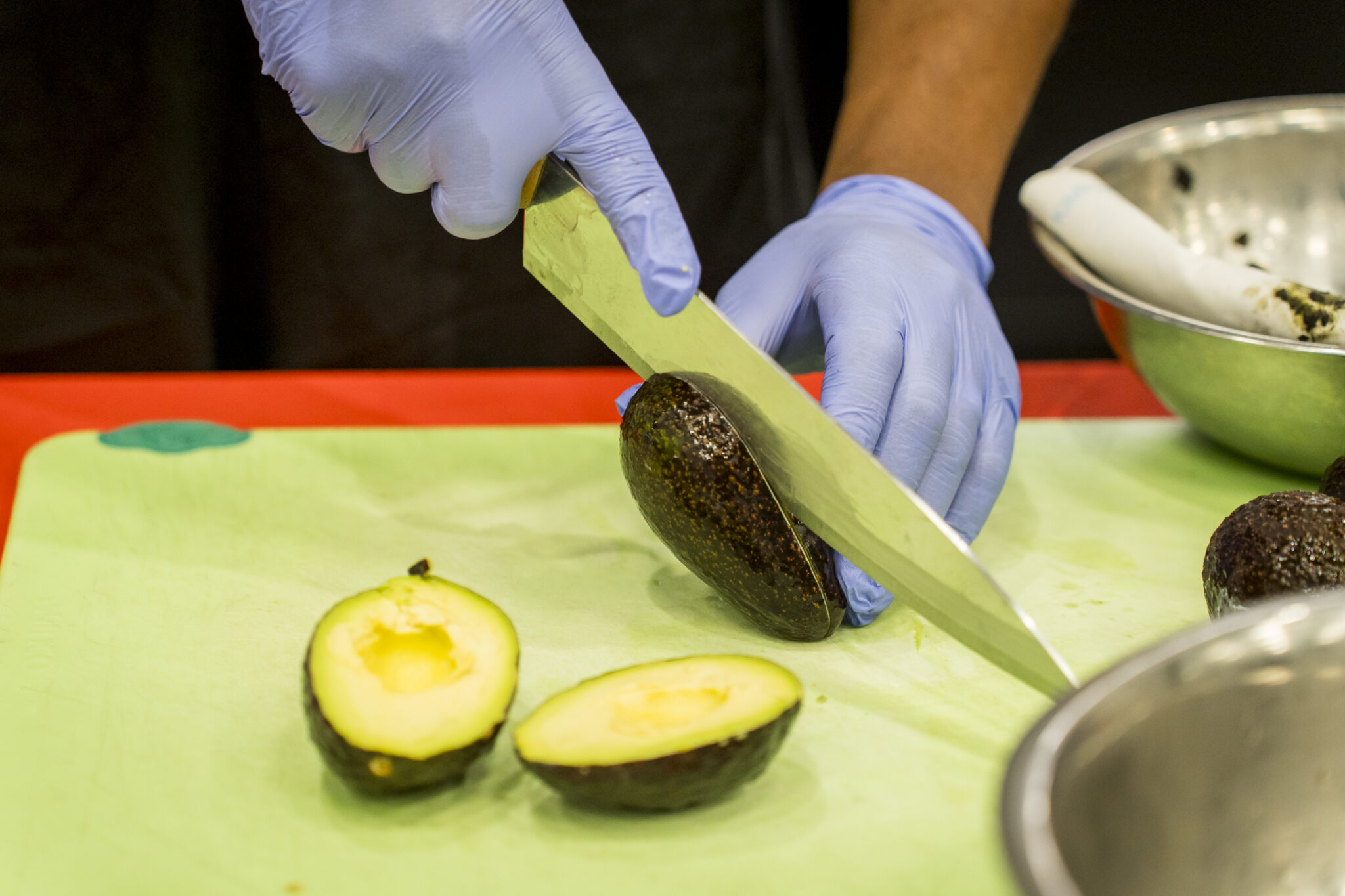 A closeup of two gloved hands holding a knife and halving an avocado on a green cutting board