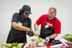 Two contestants mix avocados in a silver bowl on a table surrounded by avocados and other vegetables