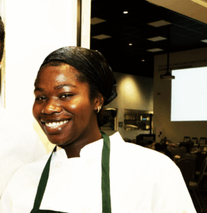 Shiffon Carrol dressed in a white chef's coat and an apron looking at the camera and smiling